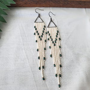 May include: A pair of long, white beaded earrings with green accents. The earrings are made of small, white beads and have a triangular shape at the top. The green accents are small, round beads that are scattered throughout the earrings.
