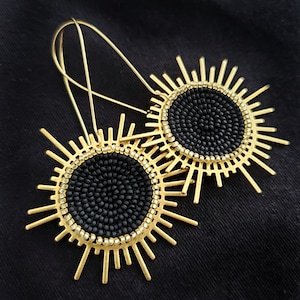 May include: A pair of gold-colored earrings with a sunburst design. The earrings feature a central black beaded circle surrounded by a gold-colored metal frame with radiating spikes. The earrings hang from gold-colored ear wires.