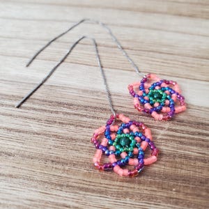 May include: A pair of threader earrings featuring geometric flower-shaped pendants crafted from small glass beads in shades of coral, purple, blue, and green. The pendants are attached to thin, silver-toned metal chains that hang from the ear.