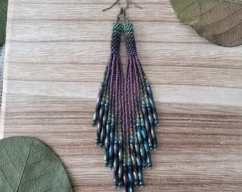 May include: Handmade beaded earrings in shades of purple, green, and blue. The earrings feature a fringe design with a gradient effect, transitioning from dark to light colors. The earrings are displayed on a wooden surface with a leaf.