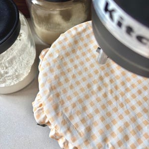 May include: A close-up shot of a kitchen scene featuring a stand mixer and jars. A fabric cover with a yellow and white checkered pattern is on the mixer. Jars with black lids are visible, one containing flour. The mixer has the word "Kitchen" on it.