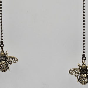 May include: Two decorative bronze-colored bee-shaped ceiling fan pull chains. Each bee pendant is intricately detailed with openwork wings and body, suspended from a dark metal ball chain against a white background.