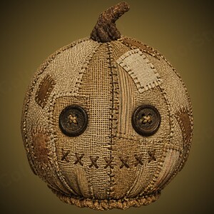 Puede incluir: Un objeto redondo con forma de calabaza hecho de tela de arpillera color canela con costuras visibles. Presenta dos botones circulares de color marrón oscuro como ojos y una boca cosida con hilo marrón en forma de X, rematado con un tallo texturizado.