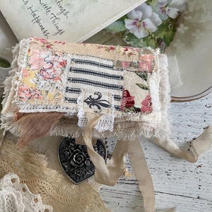 May include: A small, handmade fabric journal with a patchwork design featuring floral and striped patterns. The journal has a frayed edge and is adorned with a heart-shaped metal charm and cream-colored ribbon. The background includes a vintage book and lace.