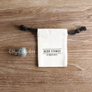 Op de afbeelding: Een witte trekkoordtas met zwarte trekkoorden en de tekst "BEER STONES BY BREW MUSE" gedrukt op de voorkant. Twee grijze bierkoelers zijn op de voorgrond.