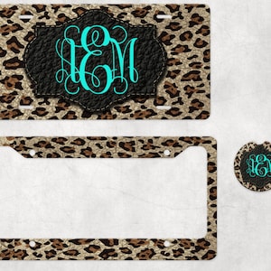 May include: A set of three leopard print car accessories featuring a license plate frame, a license plate cover, and a car air freshener. The accessories are decorated with a black leather-like patch with a monogram in turquoise.