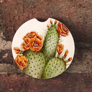 May include: A round white coaster with a green cactus and orange flowers painted on it. The coaster is made of cork and has a felt bottom.