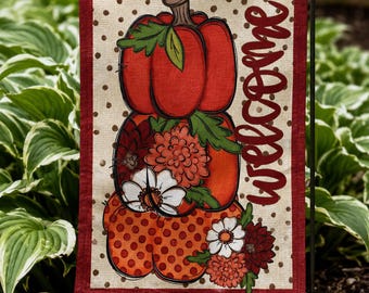 May include: A rectangular garden flag with a dark red border and a beige background featuring brown polka dots. The design displays a stack of three pumpkins decorated with orange, red, and white flowers, with the word Welcome in red script.
