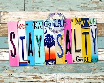 Puede incluir: Un letrero decorativo hecho de placas de matrícula, que deletrea "STAY SALTY" en varios colores. El fondo es un patrón de madera desgastada. El letrero es una pieza única de arte mural.