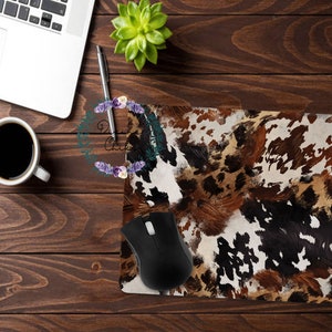 May include: A rectangular mousepad with a cowhide pattern in shades of brown, black, and white. A black computer mouse rests on the pad. The mousepad is on a wooden desk with a laptop, a cup of coffee, a pen, and a small plant.