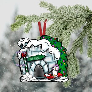Puede incluir: Un adorno navideño con un iglú blanco y azul con una puerta verde y un pingüino montando un trineo rojo. El iglú tiene un letrero que dice "TUS PALABRAS" y está decorado con un bastón de caramelo y bolas de nieve. El iglú está rodeado de ramas de pino verdes con luces rojas y verdes.