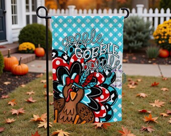 May include: A turquoise and white checkered garden flag with a cartoon turkey design. The turkey has a red bow, a feather headdress, and the words "gobble gobble gobble" in white script. The flag is displayed outdoors on a black metal stand.