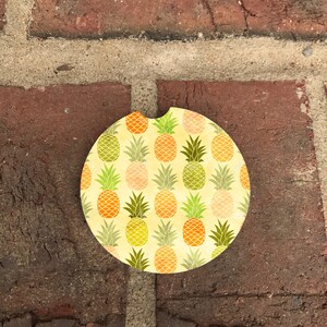 May include: A round, yellow and green coaster with a pineapple pattern. The coaster is made of a cork-like material and has a hole in the center for a beverage.