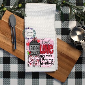 May include: White waffle-weave kitchen towel with a graphic that reads "I can't love you more than my hometown." The graphic includes a water tower, hearts, and the number 28164. The towel is displayed with wooden cutting boards, measuring cups, and tongs.