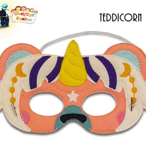 May include: A pink felt unicorn mask with a yellow horn, white wings, and purple stripes. The mask has a white star and a crescent moon with gold beads. The text "TEDDICORN" is printed above the mask.