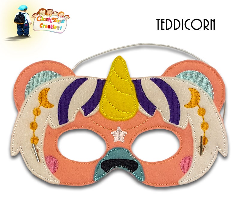May include: A pink felt unicorn mask with a yellow horn, white wings, and purple stripes. The mask has a white star and a crescent moon with gold beads. The text "TEDDICORN" is printed above the mask.