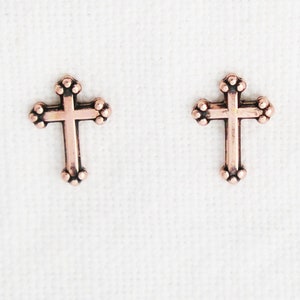 Pode incluir: Um par de pequenos brincos de cruz de cor cobre. As cruzes têm um design simples e tradicional com uma superfície texturizada.