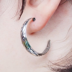 May include: A silver-toned hoop earring with an ornate floral and swirl design. The earring is curved and worn in the ear, showcasing the intricate details of the metalwork. The design includes a flower and leaf pattern.