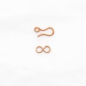 May include: Two copper-colored jewelry clasps. One is a hook shape, and the other is an infinity symbol. The clasps are small and delicate, likely for necklaces or bracelets. They are set against a white background.