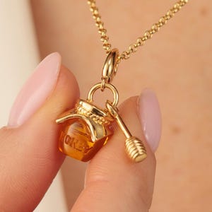May include: A gold-toned necklace featuring a small, translucent amber-colored honey pot charm with the word HONEY embossed on the front, accompanied by a matching gold-toned honey dipper charm, both suspended from a delicate gold-toned chain.