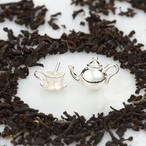 May include: A pair of silver earrings shaped like a teapot and a teacup. The earrings are set against a background of loose leaf tea.