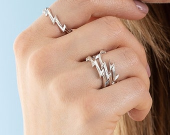 May include: A set of silver-toned rings featuring a repeating lightning bolt pattern. One ring is worn on the index finger, while two stacked rings with the same jagged, angular lightning bolt design are worn on the ring finger of the same hand.