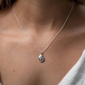 Puede incluir: Un collar con colgante de plata con un peque&ntilde;o colgante ovalado. El colgante tiene un dise&ntilde;o detallado y cuelga de una delicada cadena. El collar se lleva alrededor del cuello.