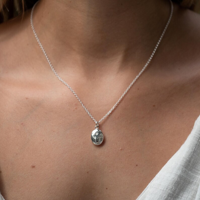 Puede incluir: Un collar con colgante de plata con un peque&ntilde;o colgante ovalado. El colgante tiene un dise&ntilde;o detallado y cuelga de una delicada cadena. El collar se lleva alrededor del cuello.