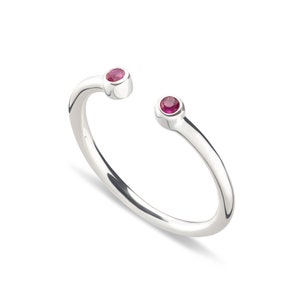 May include: A polished silver open-style ring featuring two small, round, faceted deep pink rubies set at each end of the band. The band has a smooth, reflective finish and a simple, minimalist construction with an open gap between the two gemstones.