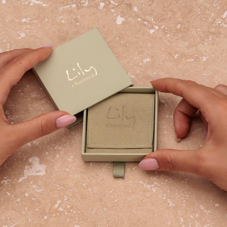 Puede incluir: Una caja de joyer&iacute;a verde claro con el texto "Lily charmed" en dorado. La caja est&aacute; abierta, revelando un interior beige y una peque&ntilde;a bolsa. La caja est&aacute; abierta por dos manos con esmalte de u&ntilde;as rosa.