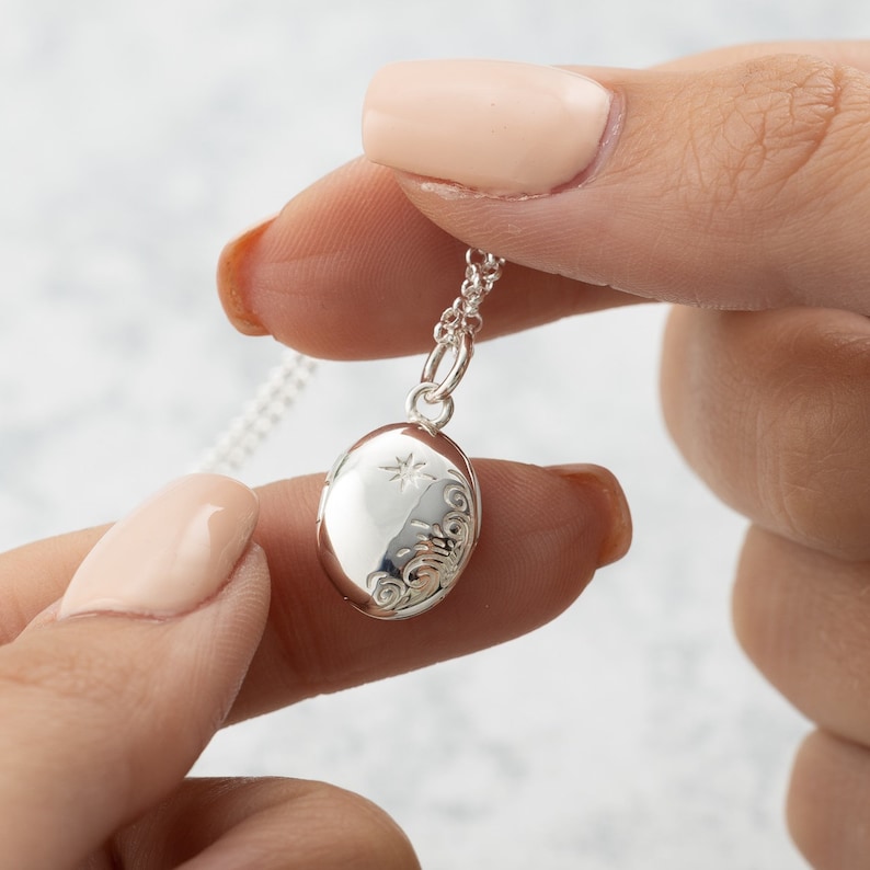 Puede incluir: Un collar con un colgante ovalado de plata y una fina cadena. El medall&oacute;n presenta un dise&ntilde;o de estrella y adornos decorativos. El collar se sostiene entre dos dedos.