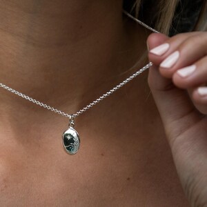 Puede incluir: Un collar de plata con un peque&ntilde;o colgante ovalado. El colgante presenta un dise&ntilde;o de sol, estrellas y patrones arremolinados en color verde azulado oscuro. El collar est&aacute; siendo sostenido por una persona.