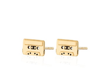 May include: A pair of gold-toned stud earrings shaped like miniature cassette tapes. Each earring features a rectangular frame with a central horizontal bar, two circular reel cutouts, and small black detailing to mimic the look of a vintage tape.
