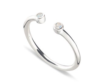 May include: A silver open-style ring featuring two small, round, iridescent moonstone cabochons set at each end of the band. The polished metal band has a smooth, reflective surface and a gap between the two stones, creating a bypass design.
