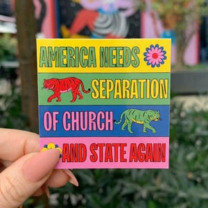 May include: A square sticker with four horizontal color bands. The yellow top band reads AMERICA NEEDS with a pink flower. The green second band shows a red tiger and the word SEPARATION. The blue third band shows a green tiger and OF CHURCH. The pink bottom band reads AND STATE AGAIN.