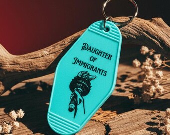 Könnte beinhalten: Ein türkisfarbener Schlüsselanhänger im Motel-Stil mit den Worten "DAUGHTER OF IMMIGRANTS" und einer schwarzen Illustration der Freiheitsstatue. Der Schlüsselanhänger ist an einem silbernen Metallring befestigt. Der Hintergrund umfasst eine Holzoberfläche und einen roten Hintergrund.