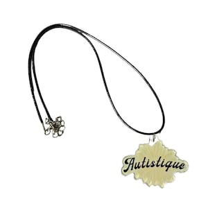 May include: A black cord necklace with a pendant. The pendant is a light yellow shape with the word "Autistique" in black script. The necklace has a silver-colored clasp.