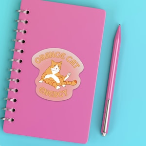 Orange Cat Energy Glossy Die Cut Vinyl Sticker on top of pink notebook with pink pen