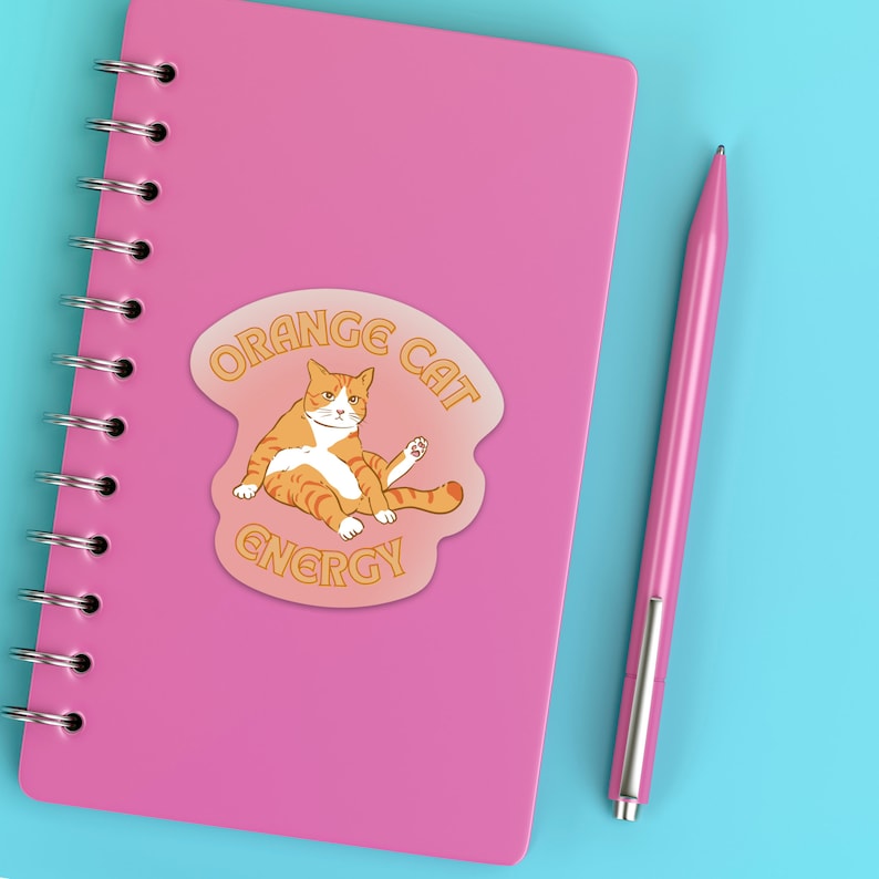 Orange Cat Energy Glossy Die Cut Vinyl Sticker on top of pink notebook with pink pen