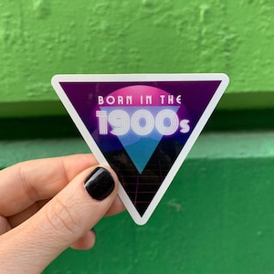 Könnte beinhalten: Ein dreieckiger Aufkleber mit einem violetten Hintergrund und einem weißen Umriss. Der Aufkleber zeigt einen rosa-blauen Farbverlaufskreis mit dem Text "Born in the 1900s" in weißen umrandeten Buchstaben.