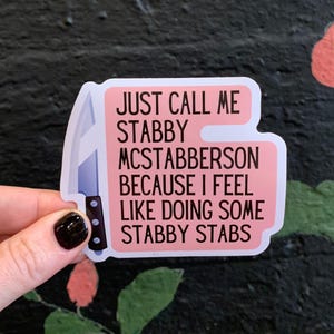May include: A pink sticker with the text "JUST CALL ME STABBY MCSTABBERSON BECAUSE I FEEL LIKE DOING SOME STABBY STABS" and a knife illustration. The sticker is held by a hand with a black painted nail.