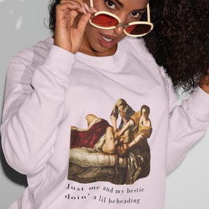 May include: A light pink sweatshirt featuring a Renaissance-style painting of a beheading scene. The text "Just me and my bestie doin' a lil beheading" is printed below the image in a handwritten style font.