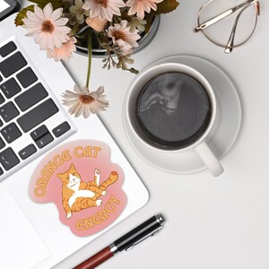 Orange Cat Energy Glossy Die Cut Vinyl Sticker on top of laptop surrounded by flower vase, coffee and eyeglass