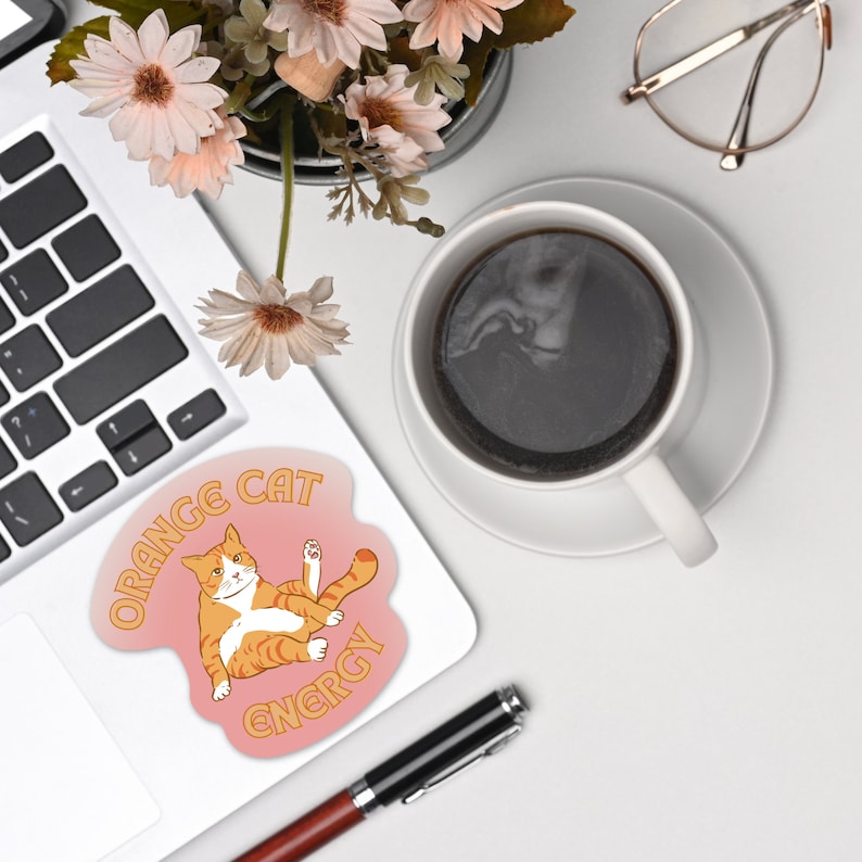 Orange Cat Energy Glossy Die Cut Vinyl Sticker on top of laptop surrounded by flower vase, coffee and eyeglass