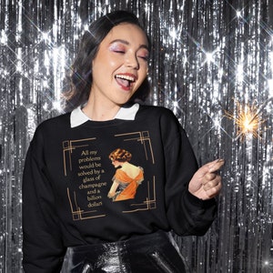 May include: Black sweatshirt with a white collar featuring a vintage illustration and text. The illustration depicts a woman in an orange dress. The text reads: "All my problems would be solved by a glass of champagne and a billion dollars."