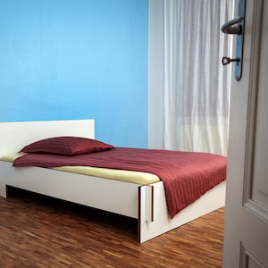 Könnte beinhalten: Ein weißes Bettgestell mit einer rot-weiß gestreiften Bettdecke und zwei Kissen. Das Bett steht in einem Raum mit blauen Wänden und einer weißen Tür.