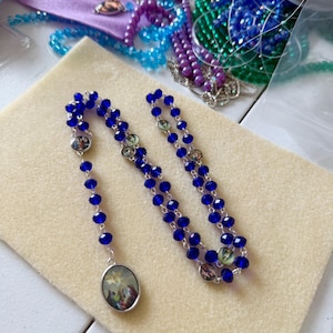 Pu&ograve; includere: Un rosario fatto a mano con perline blu vibranti e dettagli argentati. Il rosario presenta immagini religiose ovali ed &egrave; esposto su una superficie beige. Altri materiali per l'artigianato, tra cui perline e filo, sono visibili sullo sfondo.