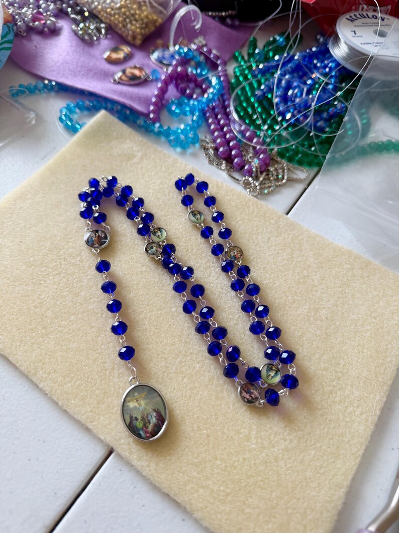 Pu&ograve; includere: Un rosario fatto a mano con perline blu vibranti e dettagli argentati. Il rosario presenta immagini religiose ovali ed &egrave; esposto su una superficie beige. Altri materiali per l'artigianato, tra cui perline e filo, sono visibili sullo sfondo.