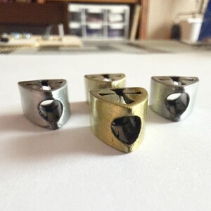 May include: Four metal thimble-shaped objects. Three are silver, and one is gold-colored. Each has a triangular top with a small hole and a heart-shaped opening on the side. The objects are arranged on a white surface.