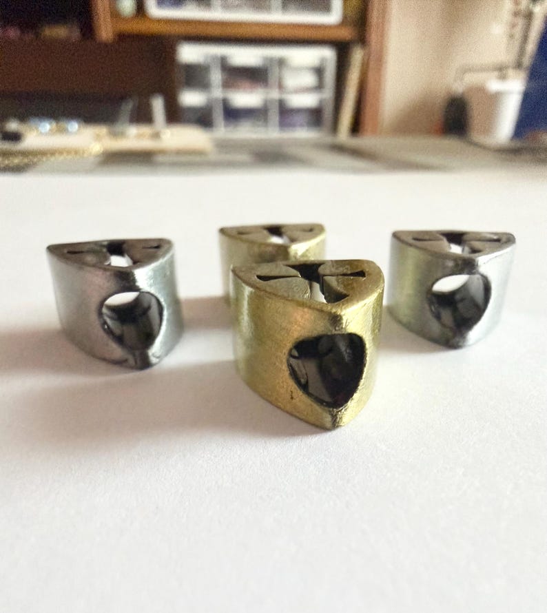 May include: Four metal thimble-shaped objects. Three are silver, and one is gold-colored. Each has a triangular top with a small hole and a heart-shaped opening on the side. The objects are arranged on a white surface.